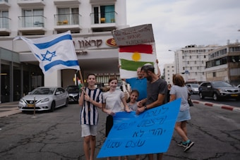 A group of people standing on a street holding flags and signs. One person holds an Israeli flag, while another holds a sign with a Kurdish flag. They appear to be in front of a hotel with cars parked along the street.