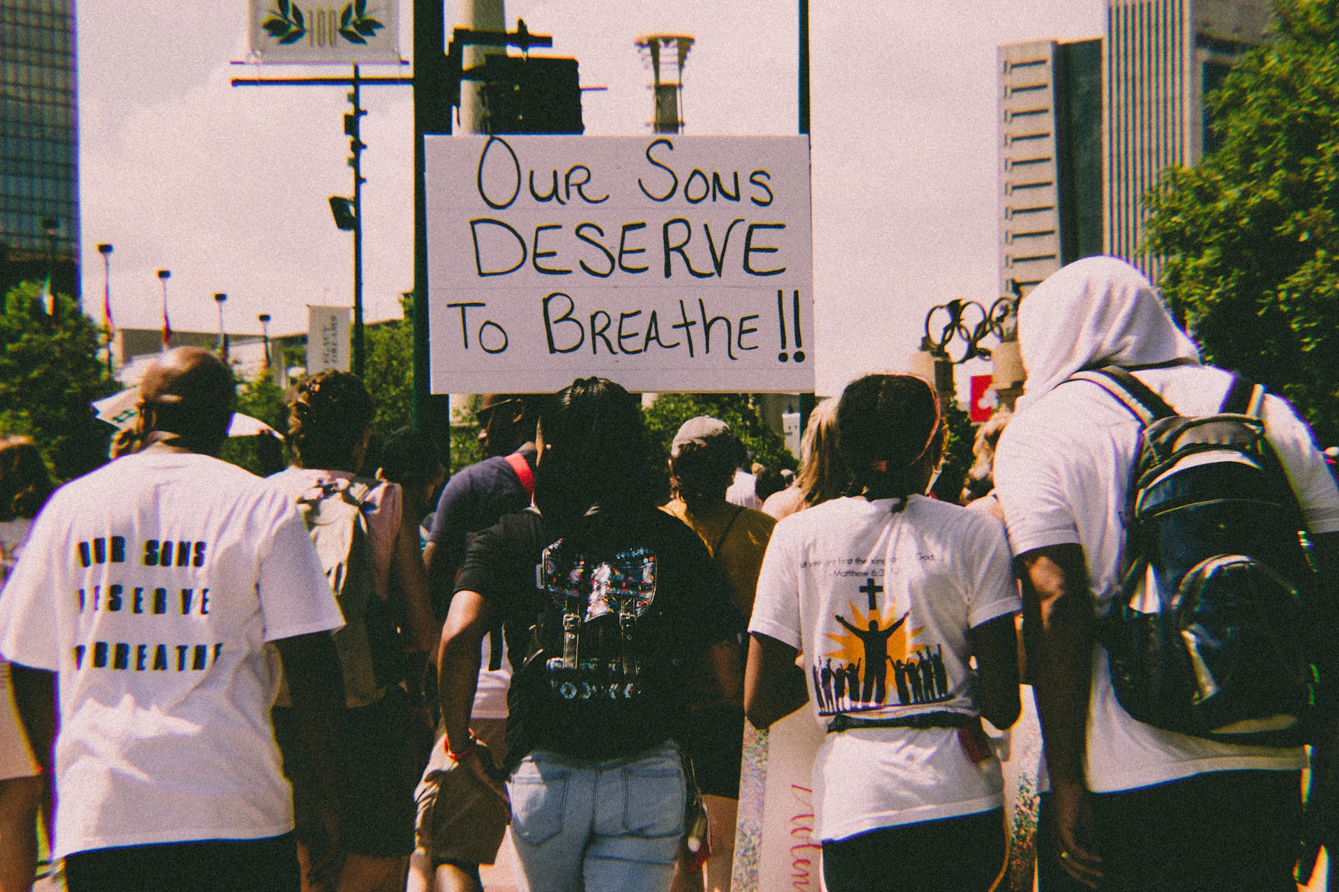 A group of people are walking in a protest holding a sign that reads 'Our sons deserve to breathe.' The crowd appears diverse, with individuals wearing casual clothing, some with backpacks. The background includes tall buildings, lush green trees, and urban street elements.