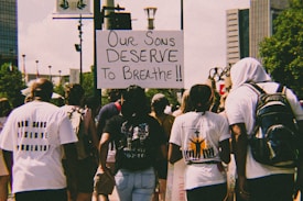 A group of people are walking in a protest holding a sign that reads 'Our sons deserve to breathe.' The crowd appears diverse, with individuals wearing casual clothing, some with backpacks. The background includes tall buildings, lush green trees, and urban street elements.