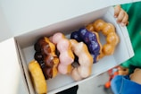 Overhead shot of hands reaching into a box filled with various uniquely topped Smithfield Ave Donuts.