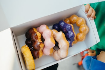 A white box contains a variety of donuts with unique bubble shapes, each topped with different colored glazes, including chocolate, pink, and purple. The open box is being held by hands, suggesting a casual or friendly sharing moment.