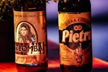 Two bottles of beer are prominently displayed, each with distinct labels. The left bottle, labeled 'Colomba,' features an illustration of a woman's face with a hood, surrounded by leaves. The right bottle is labeled 'Pietra' and displays a rustic design with text emphasizing its Corsican origin. The background includes a faintly visible plant to the left and a warm-colored surface.