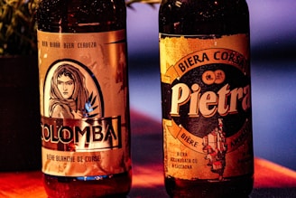 Two bottles of beer are prominently displayed, each with distinct labels. The left bottle, labeled 'Colomba,' features an illustration of a woman's face with a hood, surrounded by leaves. The right bottle is labeled 'Pietra' and displays a rustic design with text emphasizing its Corsican origin. The background includes a faintly visible plant to the left and a warm-colored surface.