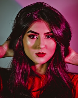 A person with long dark hair, styled with waves, poses with hands touching their head. They have vibrant red lipstick, pronounced makeup with strong eyeliner and eyebrows, and a softly illuminated face. The background is a gradient of pink and red tones, which complements the overall color scheme.