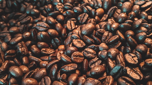 A close-up view of numerous roasted coffee beans, varying in shades from deep brown to a lighter, oily sheen. The texture appears rich and inviting, capturing the essence of freshly roasted coffee.