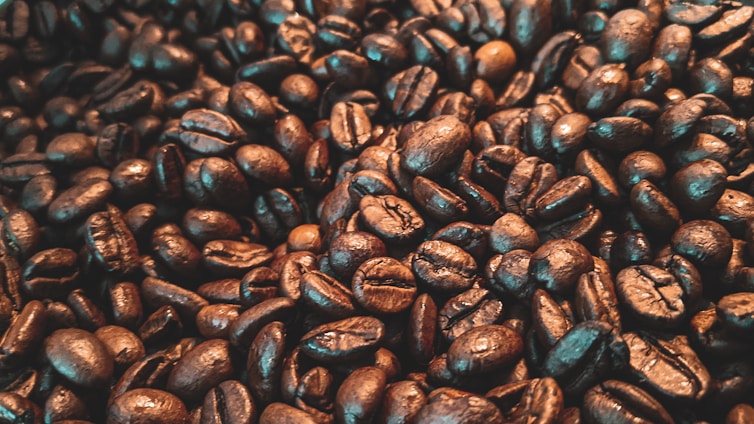 A close-up view of numerous roasted coffee beans, varying in shades from deep brown to a lighter, oily sheen. The texture appears rich and inviting, capturing the essence of freshly roasted coffee.