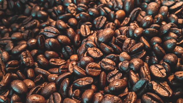 A close-up view of numerous roasted coffee beans, varying in shades from deep brown to a lighter, oily sheen. The texture appears rich and inviting, capturing the essence of freshly roasted coffee.