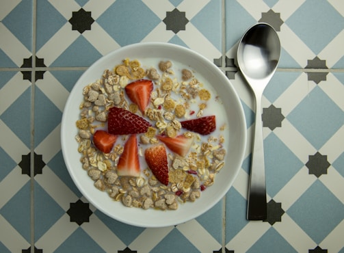 A bowl of cereal with milk, topped with sliced strawberries, is placed on a decorative blue and white tiled surface. A shiny silver spoon rests beside the bowl.