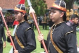 Two people wearing traditional black uniforms with gold accents are holding ceremonial staffs. They appear to be part of a cultural or historical event, engaging with others in the background who are similarly dressed.