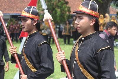 Two people wearing traditional black uniforms with gold accents are holding ceremonial staffs. They appear to be part of a cultural or historical event, engaging with others in the background who are similarly dressed.