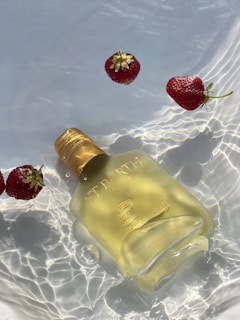 A golden bottle labeled 'ST BARTH' floats elegantly in clear, rippling water alongside several fresh strawberries. Sunlight creates beautiful reflections and patterns on the water surface, enhancing the serene and refreshing atmosphere.