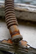 A close-up of a rusted spring with a circular metal piece at its end, resting on a surface with peeling white paint. The focus is on the corroded texture of the spring, highlighting the orange and brown rust.