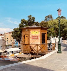 A wooden taxi booth with a sign for 'Taxi' and 'Casino di Venezia' sits beside a canal. Two water taxis are moored next to it. The setting includes a stone walkway, green street lamps, and lush trees in the background. A bridge is visible further along the canal.