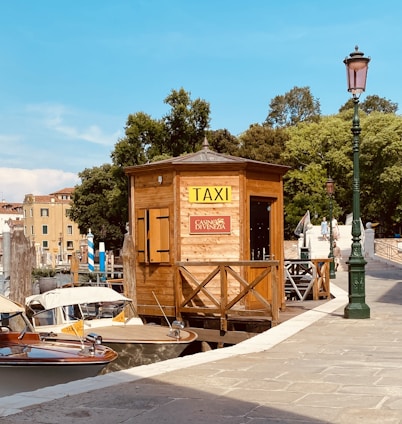 A wooden taxi booth with a sign for 'Taxi' and 'Casino di Venezia' sits beside a canal. Two water taxis are moored next to it. The setting includes a stone walkway, green street lamps, and lush trees in the background. A bridge is visible further along the canal.