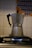 A metal stovetop espresso maker sits on a lit burner of a gas stove. The background features a tiled wall with a decorative pattern of yellow and blue diamond shapes.