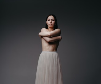 A person stands with their arms crossed over their chest, eyes closed, conveying a sense of introspection or serenity. The background is a plain, dark gradient which emphasizes the subject whose lower body is covered in a flowing, light-colored skirt.