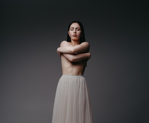 A person stands with their arms crossed over their chest, eyes closed, conveying a sense of introspection or serenity. The background is a plain, dark gradient which emphasizes the subject whose lower body is covered in a flowing, light-colored skirt.