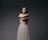 A person stands with their arms crossed over their chest, eyes closed, conveying a sense of introspection or serenity. The background is a plain, dark gradient which emphasizes the subject whose lower body is covered in a flowing, light-colored skirt.