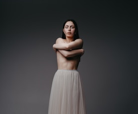 A person stands with their arms crossed over their chest, eyes closed, conveying a sense of introspection or serenity. The background is a plain, dark gradient which emphasizes the subject whose lower body is covered in a flowing, light-colored skirt.
