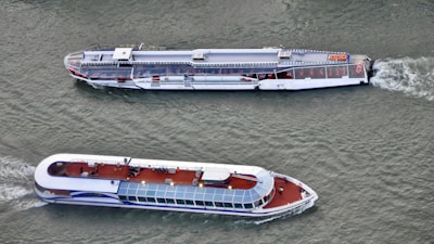 Two modern passenger boats are cruising on a body of water. Both boats have sleek designs with open viewing decks and have a white and blue color scheme. The vessels are moving parallel to each other, and there are visible ripples in the water created by their motion.