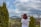 A person with red hair and a white shirt is using a camera with a large lens, overlooking a scenic landscape with distant mountains. The foreground features a decorative iron fence and neatly trimmed bushes. The sky is partly cloudy, creating a serene atmosphere.
