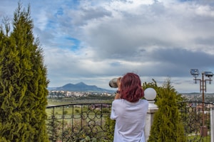 A person with red hair and a white shirt is using a camera with a large lens, overlooking a scenic landscape with distant mountains. The foreground features a decorative iron fence and neatly trimmed bushes. The sky is partly cloudy, creating a serene atmosphere.
