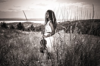 A person stands in a grassy field holding a violin, with long curly hair and wearing a sleeveless dress. The background includes a distant view of a city, a body of water, and the sky partially clouded. The image is rendered in a sepia tone, giving it a vintage feel.