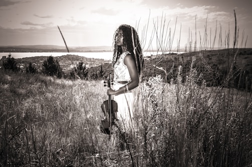 A person stands in a grassy field holding a violin, with long curly hair and wearing a sleeveless dress. The background includes a distant view of a city, a body of water, and the sky partially clouded. The image is rendered in a sepia tone, giving it a vintage feel.