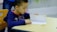 A young child is seated at a desk, focused on a worksheet with numbers and arrows. The child is wearing a blue hoodie and holding a pencil. The setting appears to be a classroom, indicated by the desk and chair.