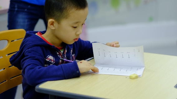 A young child is seated at a desk, focused on a worksheet with numbers and arrows. The child is wearing a blue hoodie and holding a pencil. The setting appears to be a classroom, indicated by the desk and chair.