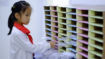 A young girl wearing a white shirt with a red scarf stands in front of a wall of mail slots. She appears to be sorting or retrieving items from the slots which are organized in multiple rows and columns.