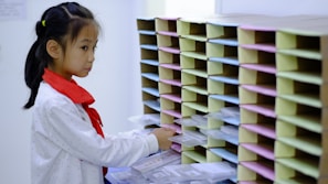 A young girl wearing a white shirt with a red scarf stands in front of a wall of mail slots. She appears to be sorting or retrieving items from the slots which are organized in multiple rows and columns.