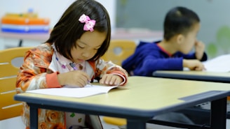 a little girl sitting at a table writing on a piece of paper