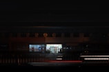 A brightly lit convenience store is surrounded by a dimly lit urban environment. The store's signage illuminates the surroundings, contrasting with the darkness. A few parked cars and bicycles can be seen nearby.