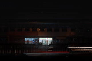 A brightly lit convenience store is surrounded by a dimly lit urban environment. The store's signage illuminates the surroundings, contrasting with the darkness. A few parked cars and bicycles can be seen nearby.
