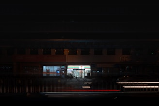 A brightly lit convenience store is surrounded by a dimly lit urban environment. The store's signage illuminates the surroundings, contrasting with the darkness. A few parked cars and bicycles can be seen nearby.