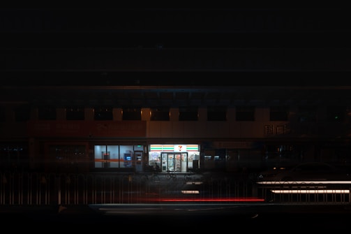 A brightly lit convenience store is surrounded by a dimly lit urban environment. The store's signage illuminates the surroundings, contrasting with the darkness. A few parked cars and bicycles can be seen nearby.