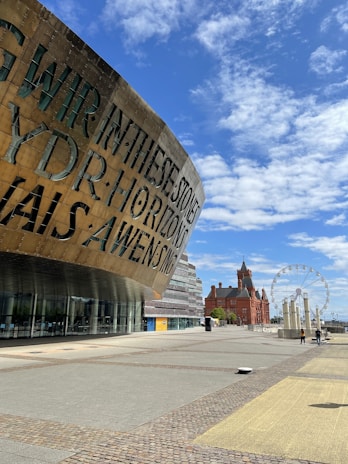 A modern architectural structure with large, metallic lettering in two languages stands prominently, next to a spacious, open plaza. In the background, a ferris wheel and a historic red-brick building with pointed towers are visible under a clear blue sky with scattered clouds.