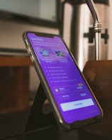 A smartphone is placed on a black stand showing a subscription page for an app. The screen displays a purple background with text about various features and pricing options, highlighting a 'Get Pro!' offer. The surrounding environment includes a blurred background and a metallic object nearby.