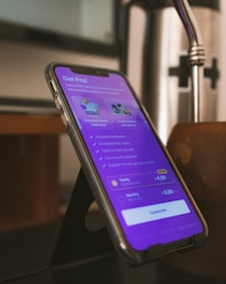 A smartphone is placed on a black stand showing a subscription page for an app. The screen displays a purple background with text about various features and pricing options, highlighting a 'Get Pro!' offer. The surrounding environment includes a blurred background and a metallic object nearby.