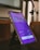 A smartphone is placed on a black stand showing a subscription page for an app. The screen displays a purple background with text about various features and pricing options, highlighting a 'Get Pro!' offer. The surrounding environment includes a blurred background and a metallic object nearby.
