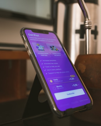 A smartphone is placed on a black stand showing a subscription page for an app. The screen displays a purple background with text about various features and pricing options, highlighting a 'Get Pro!' offer. The surrounding environment includes a blurred background and a metallic object nearby.