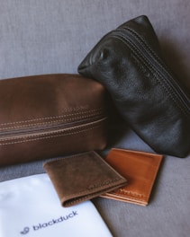 The image features two leather pouches, one dark brown and one black, positioned on a fabric surface. In front of them are two leather wallets, one brown and the other tan, along with a branded white cloth bag labeled 'blackduck'. The items appear to be high-quality leather goods.