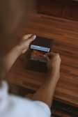A person is holding an open wallet over a wooden surface, revealing a few cards including an ID and a credit card with contactless payment symbol. The person has light purple nail polish.