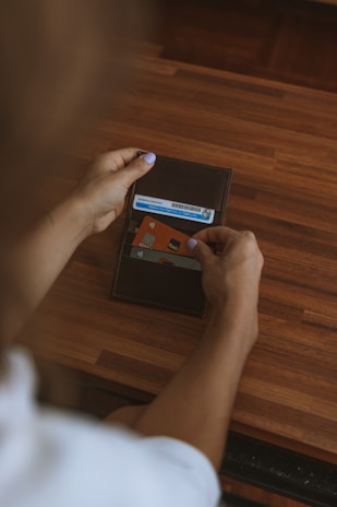 A person is holding an open wallet over a wooden surface, revealing a few cards including an ID and a credit card with contactless payment symbol. The person has light purple nail polish.