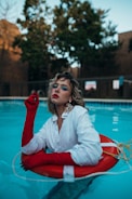 A person wearing a white button-up shirt and bright red gloves is leaning in a pool while holding a life preserver. The individual has curly blonde hair and is adorned with bold blue eye makeup and colorful earrings. Surrounding the pool are trees and a building in the background.