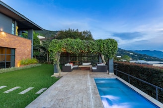 A modern backyard featuring a rectangular swimming pool bordered by stone tiles. To one side, an outdoor seating area is sheltered by a pergola covered with lush green vines. Adjacent is a contemporary house with a mix of stone and dark paneling. The garden is well-manicured with green grass, and the landscape offers a scenic view of distant hills and an overcast sky.