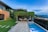 A modern backyard featuring a rectangular swimming pool bordered by stone tiles. To one side, an outdoor seating area is sheltered by a pergola covered with lush green vines. Adjacent is a contemporary house with a mix of stone and dark paneling. The garden is well-manicured with green grass, and the landscape offers a scenic view of distant hills and an overcast sky.
