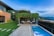 A modern backyard featuring a rectangular swimming pool bordered by stone tiles. To one side, an outdoor seating area is sheltered by a pergola covered with lush green vines. Adjacent is a contemporary house with a mix of stone and dark paneling. The garden is well-manicured with green grass, and the landscape offers a scenic view of distant hills and an overcast sky.