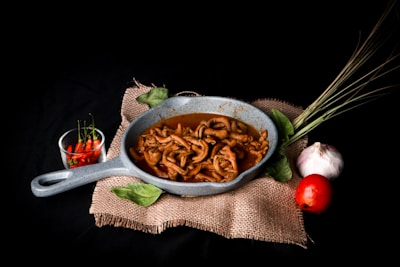 A rustic-looking grey skillet filled with cooked food, likely meat, resting on a piece of burlap cloth. Surrounding the skillet are two green leaves, a bunch of lemongrass, a white garlic bulb, and a red tomato. To the left of the skillet, there is a small glass bowl containing red chili peppers.